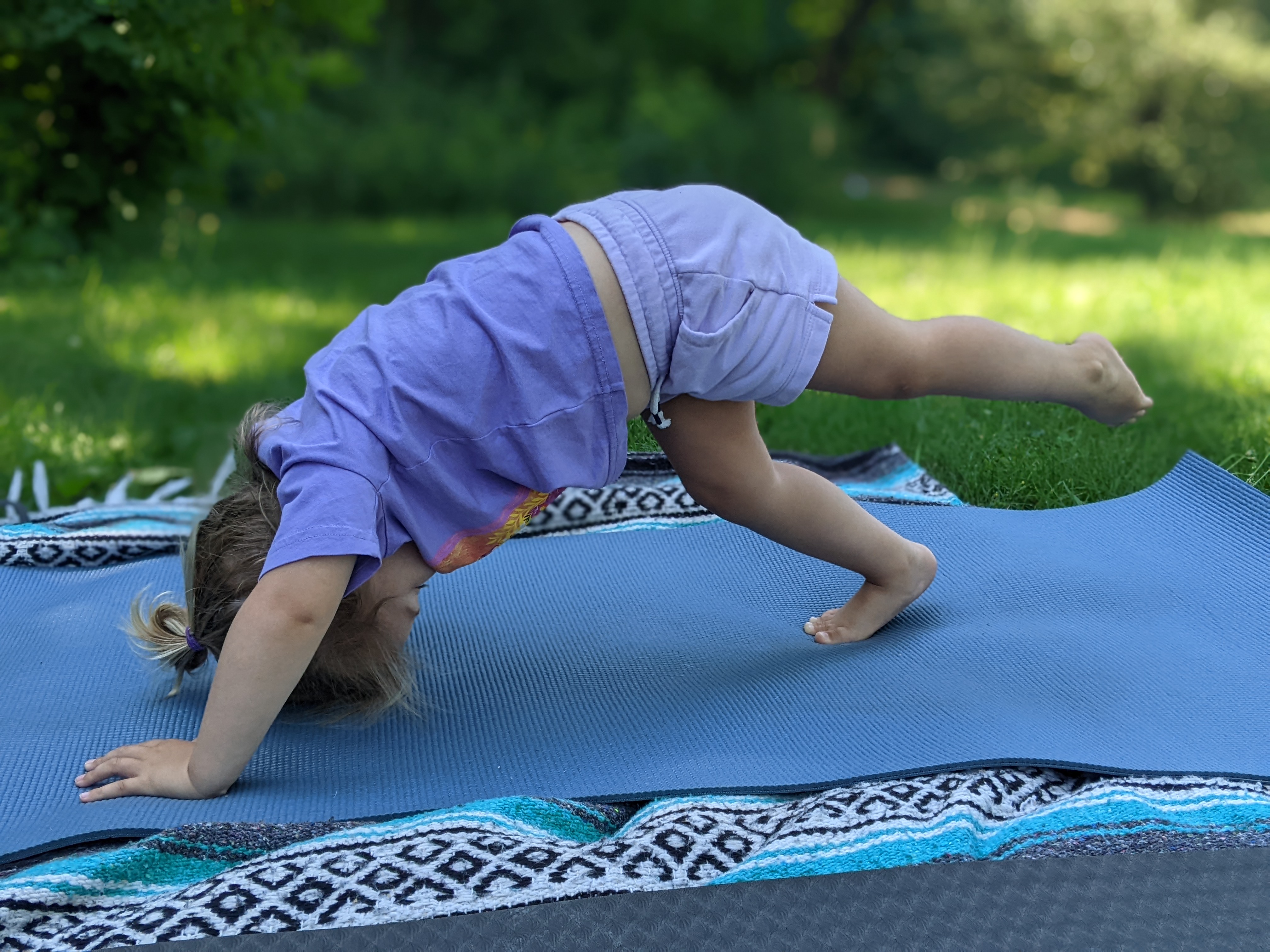 A toddler balancing on 2 hands and one foot, on a yoga mat laying in the grass with trees in the background.